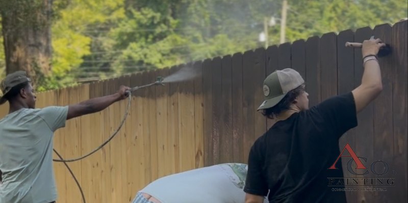 ACo. Painting crew spraying and back-brushing stain on a pressure-treated wood privacy fence in Meridian, MS, with raw pickets still visible on the left